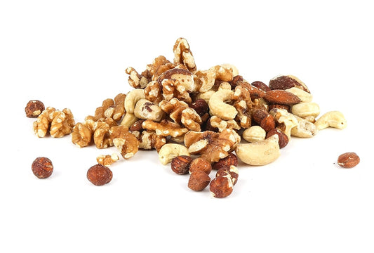 A pile of assorted nuts including walnuts, cashews, hazelnuts, and almonds is spread on a white background. The nuts are unroasted and some pieces are slightly broken, giving a raw and natural appearance.